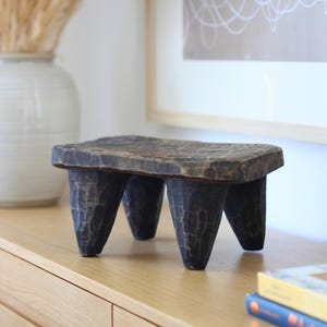 May include: A small, rectangular, dark brown wooden stool featuring a hand-carved, textured surface and four thick, tapered legs. The stool has a rustic, weathered appearance with visible chisel marks across the top and the legs, showcasing a solid, sturdy construction with a low-profile design.
