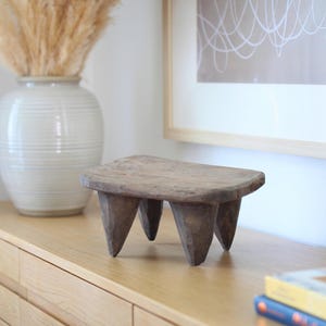 May include: A small, low-profile wooden stool featuring a rectangular top with rounded edges and four thick, hand-carved legs. The stool is crafted from dark, weathered wood with a visible grain and a rough, textured surface finish throughout.