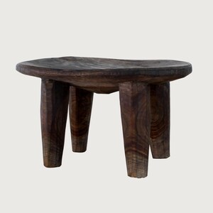 Puede incluir: Un pequeño taburete de madera marrón oscuro con una parte superior redondeada y cuatro patas robustas. La veta de la madera es visible, lo que le da un aspecto rústico. El taburete está diseñado para sentarse o como elemento decorativo.