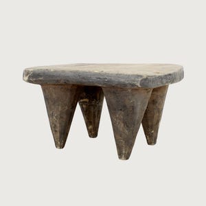 May include: A rustic, three-legged wooden stool with a triangular top. The stool is made of dark, weathered wood, with a natural, aged appearance. The legs are conical, and the top is roughly hewn, showcasing the wood's grain and texture. A unique piece of furniture.