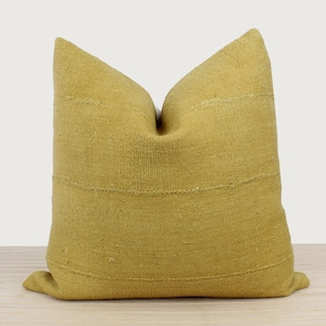 May include: A square, mustard-yellow decorative pillow with horizontal stitched lines. The pillow has a textured, woven appearance and is placed on a light-colored wooden surface. The pillow is a home decor item.