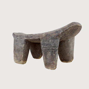 May include: A weathered, gray wooden stool with a curved seat and three cylindrical legs. The stool has a rustic appearance, with visible wood grain and a worn texture. The design is simple and functional, suitable for seating or display.
