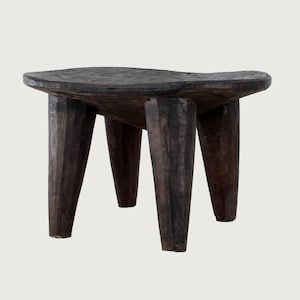 Puede incluir: Un taburete de madera tallada a mano, de color marrón oscuro, con una parte superior redondeada y cuatro patas cónicas y robustas. La veta de la madera es visible, lo que añade textura al diseño rústico. Una pieza de mobiliario única.