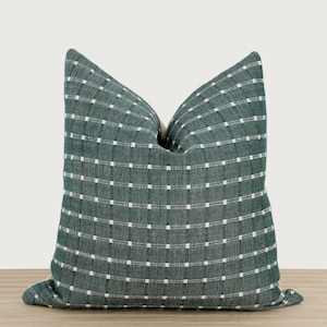 May include: A square throw pillow featuring a deep forest green fabric with a grid pattern formed by thin, vertical and horizontal white dashed lines. The pillow has a textured, woven appearance with a visible crease at the top center.
