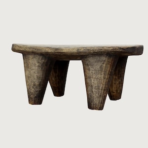 May include: A small, rustic wooden stool with a rounded top and three conical legs. The stool has a weathered, textured appearance with visible wood grain, showcasing a natural, aged aesthetic. The color is a mix of browns and grays.