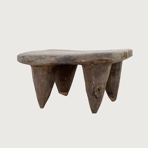 Puede incluir: Un taburete de madera de tres patas, desgastado. El taburete tiene una parte superior rectangular y tres patas cónicas. La madera es de color gris-marrón con vetas y nudos visibles, lo que sugiere edad y uso. El diseño es simple y rústico.
