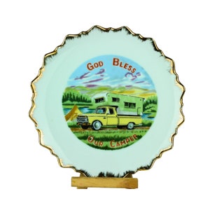 Puede incluir: Un plato decorativo con borde festoneado y ribete dorado. El plato presenta una camioneta amarilla con una caravana, una tienda de campaña y las palabras "GOD BLESS OUR CAMPER" en rojo y verde.