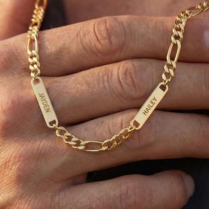 May include: A gold-colored Figaro chain necklace with two rectangular nameplates. The nameplates are engraved with the names "Jayden" and "Hailey". The necklace is displayed against a dark gray shirt.