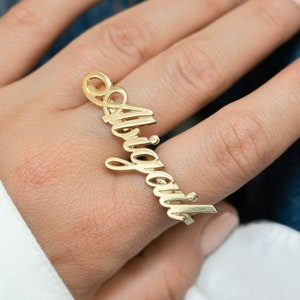 Puede incluir: Un anillo dorado con el nombre "Abigail" escrito en cursiva. El anillo se lleva en un dedo, con el nombre extendiéndose verticalmente. El anillo tiene un lazo en la parte superior y una barra en la parte inferior.