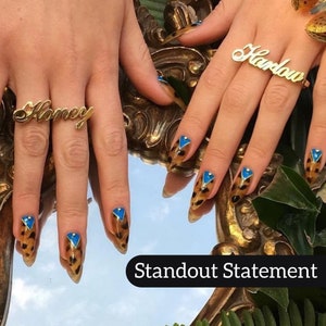 May include: Close-up of hands with long, pointed nails featuring a leopard print design with blue accents. The nails are reflected in a mirror with the text "Standout Statement" below. The hands are wearing gold rings with the words "Honey" and "Harlow" on them.