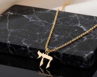 May include: A gold-colored necklace featuring the Hebrew symbol "Chai", meaning "life". The pendant hangs from a delicate chain. The necklace is displayed on a black marble surface.