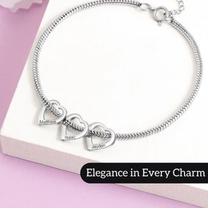 May include: Silver bracelet with three heart-shaped charms, each engraved with a name: Matthew, Isaac, and Jayden. The bracelet has a snake chain style and a lobster clasp closure. The text "Elegance in Every Charm" is displayed below the bracelet.