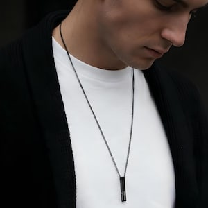 Può includere: Una collana a catena nera con un ciondolo rettangolare. Il ciondolo ha la scritta verticale "JEFFREY". La collana è indossata sopra una maglietta bianca e un cardigan nero.