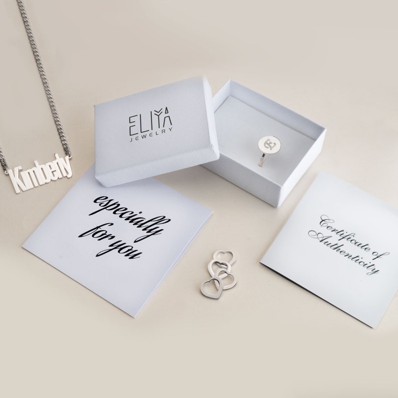 May include: A white box with the text "ELIZA JEWELRY" on the top. The box is open and a silver ring with a small letter "K" is inside. There is a white card with the text "especially for you" and a white card with the text "Certificate of Authenticity".