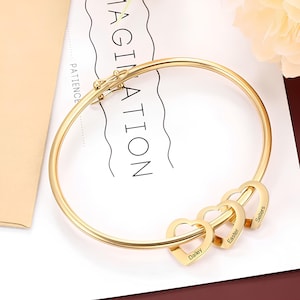 May include: Gold bangle bracelet with three heart charms. Each heart charm has a name engraved on it: Dailey, Easter, and Sellers.