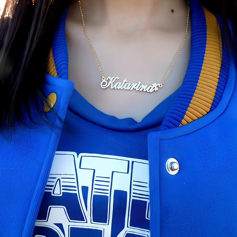 May include: A gold chain necklace with a white nameplate pendant that reads "Katarina". The necklace is worn by a person wearing a blue jacket with a yellow stripe.