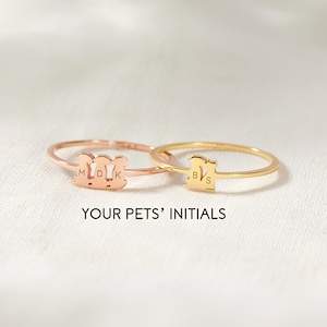 May include: Two slender rings, one rose gold and one gold, each with a pet-shaped design and initials. The rose gold ring has "MDK", the gold ring has "BVS". The text "YOUR PETS' INITIALS" is below the rings.
