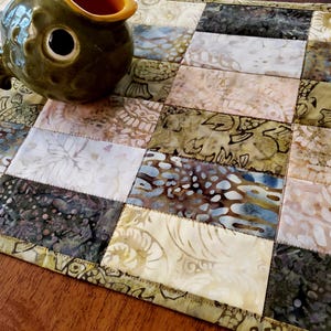 May include: A rectangular fabric table runner featuring a patchwork design of batik squares in shades of olive green, cream, brown, and blue. A ceramic fish-shaped container with a dark green exterior and orange interior sits on top.