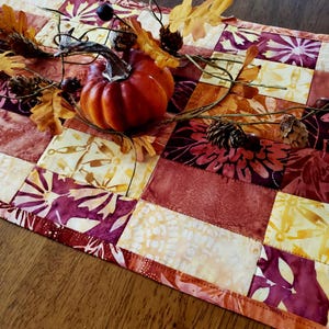 May include: A rectangular quilted table runner featuring a patchwork design of fabric squares in shades of burgundy, orange, yellow, and cream. A decorative arrangement of a small orange pumpkin, pinecones, and faux leaves rests on top.