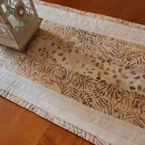 May include: A decorative table runner featuring a central panel with a brown leaf pattern on a cream background. The runner is bordered by white fabric and has a quilted design. A decorative lantern sits on the runner.