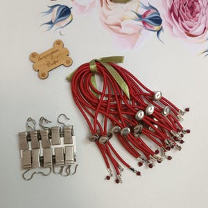 Könnte beinhalten: Ein Set roter Gummibänder für Haare mit silbernen Perlen und Metallverschlüssen. Die Haargummis sind mit einem grünen Band zusammengebunden. Das Set enthält außerdem eine Packung silberner Metallclips mit Haken.