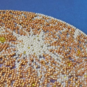 May include: Close-up of a beaded design with white, gold, and yellow beads. The beads are arranged in a circular pattern, creating a floral motif.