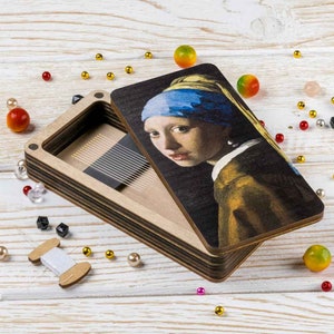 May include: A wooden box with a hinged lid featuring a reproduction of the famous "Girl with a Pearl Earring" painting. The box is open, revealing a compartment with a row of small, thin needles.