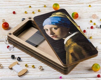 May include: A wooden box with a hinged lid featuring a reproduction of the famous "Girl with a Pearl Earring" painting. The box is open, revealing a compartment with a row of small, thin needles.