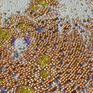 May include: Close-up of a beaded artwork with a pattern of white, gold, and yellow beads. The beads are arranged in a repeating pattern, creating a textured surface.