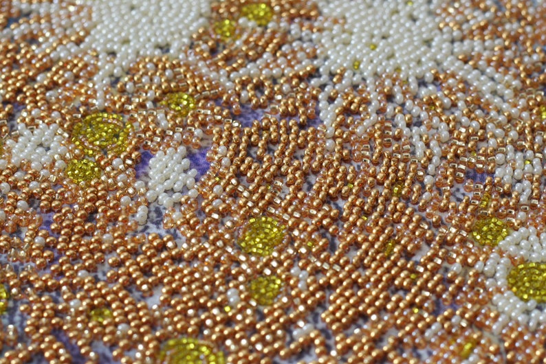 May include: Close-up of a beaded artwork with a pattern of white, gold, and yellow beads. The beads are arranged in a repeating pattern, creating a textured surface.