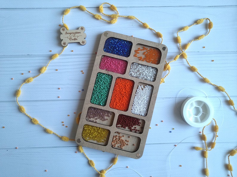 Puede incluir: Un organizador de cuentas de madera con 12 compartimentos llenos de cuentas de varios colores. El organizador est&aacute; etiquetado como "Inspiration Crafts".
