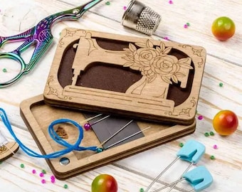Op de afbeelding: Een houten naaibox met een uitgesneden ontwerp van een naaimachine en bloemen. De doos is open en bevat blauwe draad, naalden en veiligheidsspelden.