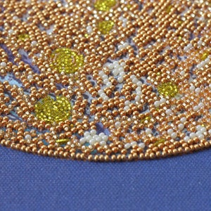 May include: Close-up of a beaded design with a circular pattern. The beads are in shades of gold, white, and yellow. The design is on a blue fabric background.