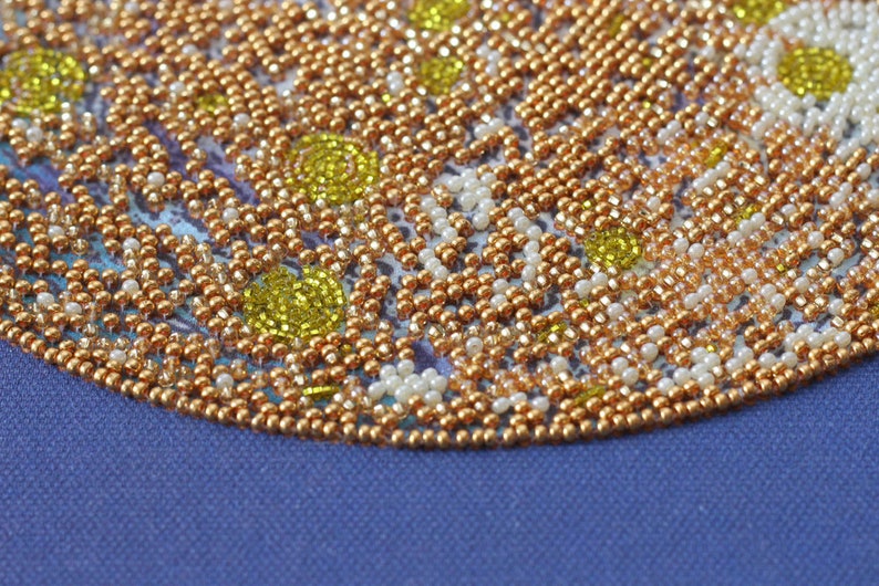 May include: Close-up of a beaded design with a circular pattern. The beads are in shades of gold, white, and yellow. The design is on a blue fabric background.