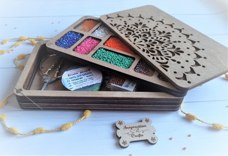 Puede incluir: Caja de madera con un dise&ntilde;o de mandala en la tapa. La caja tiene compartimentos para guardar cuentas y otros objetos peque&ntilde;os. La caja est&aacute; abierta y hay cuentas en los compartimentos. Hay un carrete de hilo y unas pinzas en la caja. La caja est&aacute; etiquetada con "Inspiration Crafts".