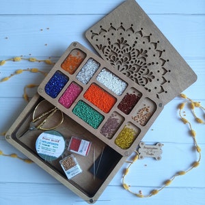 Puede incluir: Una caja de madera con un dise&ntilde;o de mandala en la tapa. La caja tiene compartimentos llenos de cuentas de diferentes colores. La caja tambi&eacute;n incluye un carrete de hilo, agujas y un peque&ntilde;o recipiente de cuentas.