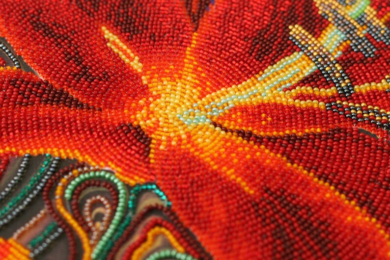 May include: Close-up of a beaded artwork depicting a red flower with yellow and blue accents. The beads are arranged in a circular pattern, creating a textured and intricate design.