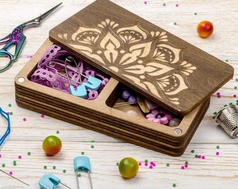 May include: A wooden box with a mandala design on the lid. The box is open and contains various sewing supplies, including pins, needles, and safety pins. The box is sitting on a white wooden surface.