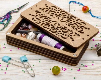 May include: A wooden box with a decorative laser-cut design on the lid. The box is open and filled with various sewing supplies, including spools of thread, buttons, and needles. The box is sitting on a white wooden surface.