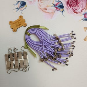 Op de afbeelding: Een verzameling lavendelkleurige elastische koorden met zilveren metalen uiteinden en kleine zwarte kralen. De koorden zijn vastgebonden met een groen lint. Ook inbegrepen zijn zilveren metalen clips en een houten label met de tekst "Inspiration for Crafts".