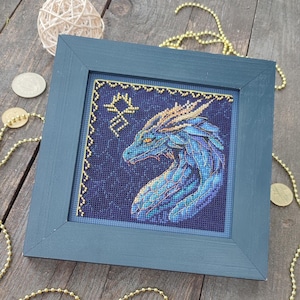 Könnte beinhalten: Eine gerahmte Kreuzstich-Stickerei eines blauen Drachen mit goldenen Akzenten. Der Drache ist im Profil dargestellt, mit dem Kopf nach links gedreht. Der Hintergrund ist dunkelblau mit einem goldenen Rand.