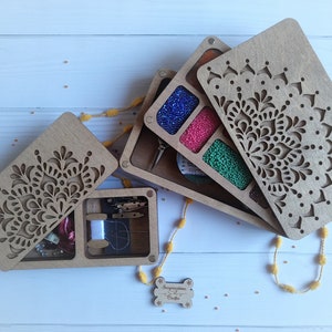 Puede incluir: Una caja de madera con un dise&ntilde;o de mandala en la tapa. La caja est&aacute; dividida en compartimentos y contiene cuentas, hilo y otros materiales de artesan&iacute;a. La caja est&aacute; abierta y los compartimentos son visibles. La caja est&aacute; sobre una superficie blanca.