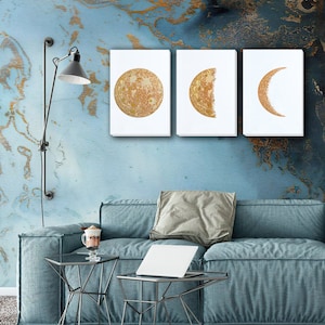 May include: Three gold-toned canvas prints of the moon in different phases. The first print shows a full moon, the second shows a half moon, and the third shows a crescent moon. The prints are hanging on a blue wall with gold accents.