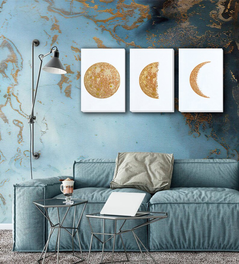 May include: Three gold-toned canvas prints of the moon in different phases. The first print shows a full moon, the second shows a half moon, and the third shows a crescent moon. The prints are hanging on a blue wall with gold accents.