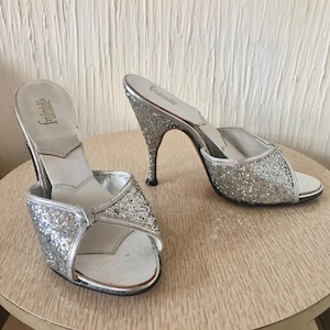 May include: A pair of silver glitter heeled sandals with a peep toe and an open back. The sandals are labeled "Frederick's of Hollywood".