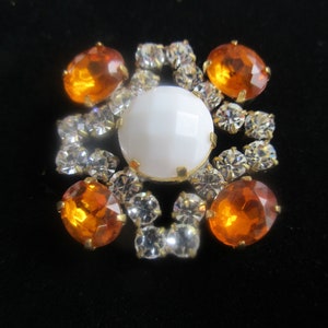 May include: A white and orange rhinestone brooch with a large white center stone surrounded by smaller orange and clear rhinestones.