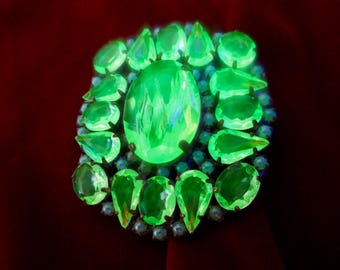 May include: A vibrant, glowing green brooch with an oval center stone and surrounding teardrop-shaped gems. The brooch is set with smaller blue-green stones, creating a striking contrast against the red background.