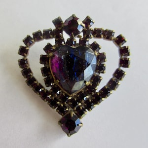 May include: A heart-shaped brooch with a large, faceted, purple glass stone in the center. The stone is surrounded by a frame of smaller, purple glass stones set in gold-toned metal.