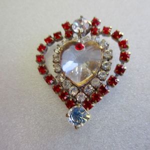 May include: A heart-shaped brooch with a large clear crystal surrounded by smaller clear and red crystals.
