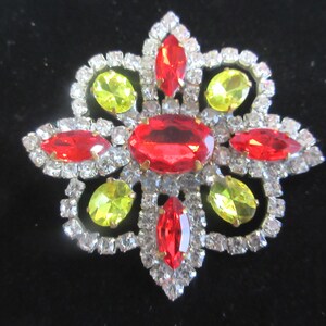 May include: A floral-shaped brooch featuring a large central oval red stone, surrounded by four marquise-cut red stones and four oval yellow stones. The entire piece is framed by small, clear, sparkling rhinestones set in a silver-tone metal.